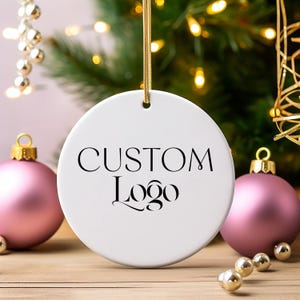 May include: White ceramic Christmas ornament with a gold string for hanging. The ornament has the text "CUSTOM Logo" printed in black.