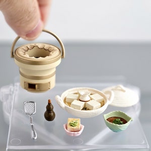 Könnte beinhalten: Ein Miniatur-japanisches Fondue-Set mit einem weißen Keramiktopf, einem kleinen braunen und weißen Herd, einem weißen Keramikdeckel, einer kleinen weißen Schüssel mit grünen und braunen Akzenten, einer kleinen rosa Schüssel mit einem weißen Tofu-Quadrat, einer kleinen braunen Kalebasse und einem silbernen Spatel.
