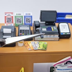 May include: Miniature cash register set with a receipt printer, card reader, and scanner. Includes toy credit cards and QR code signs. The set is on a brown surface.