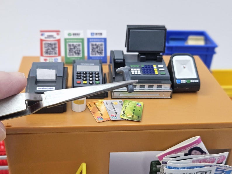 May include: Miniature cash register set with a receipt printer, card reader, and scanner. Includes toy credit cards and QR code signs. The set is on a brown surface.