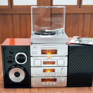 Könnte beinhalten: Miniatur-Stereoanlage in Silber mit Plattenspieler, Verstärker und Lautsprechern. Das System umfasst eine schwarze Schallplatte, eine transparente Kunststoffabdeckung und ein kleines Albumcover. Die Lautsprecher sind schwarz mit silbernen Akzenten und einem roten Oberteil.