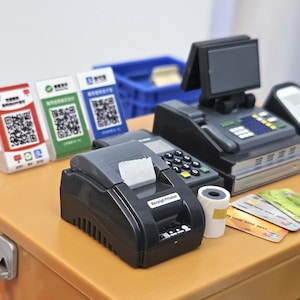 May include: A miniature cash register setup with a receipt printer, credit cards, and QR code payment displays. The scene includes a cash register with a small screen, a payment terminal, and a roll of receipt paper.