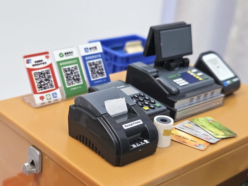 May include: A miniature cash register setup with a receipt printer, credit cards, and QR code payment displays. The scene includes a cash register with a small screen, a payment terminal, and a roll of receipt paper.