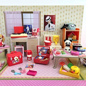 Op de afbeelding: Een miniatuur poppenhuis scène met diverse rode, witte en gele accessoires. Inclusief een pop, een platenspeler, een rode koffer en een miniatuur panda beer. De scène speelt zich af in een kamer met een raam en een poster.