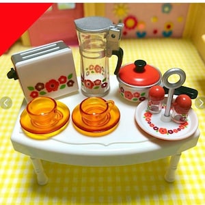 Puede incluir: Una mesa blanca con una tetera de diseño floral blanco y rojo, una tostadora de diseño floral blanco y rojo, una olla de diseño floral blanco y rojo, dos tazas de té y platillos naranjas, y un juego de salero y pimentero de diseño floral blanco y rojo.