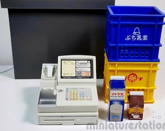 May include: A white cash register with a black and silver scanner, a blue and yellow stackable crate with the text "ふち乳業" in white, and two cartons of milk, one with the text "ふち牛乳" and the other with the text "チョコオレ" in Japanese.