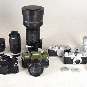 Könnte beinhalten: Eine Sammlung von Vintage-Kameras und Objektiven. Das Bild zeigt mehrere schwarz-silberne Kameras sowie verschiedene Objektive unterschiedlicher Größe. Die Marke Canon ist auf einigen Kameras sichtbar. Die Objektive sind schwarz mit silbernen Akzenten.