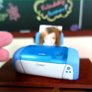 Op de afbeelding: Een blauwe en witte fotoprinter met een foto van een persoon op de achtergrond. De printer heeft een aan/uit-knop en twee andere knoppen aan de voorkant.