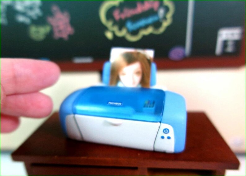 Op de afbeelding: Een blauwe en witte fotoprinter met een foto van een persoon op de achtergrond. De printer heeft een aan/uit-knop en twee andere knoppen aan de voorkant.