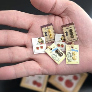 Może przedstawiać: Kolekcja małych guzików i kolczyków w stylu vintage. Guziki są złote i czarne, a kolczyki w kształcie truskawek. Karty zawierają ilustracje i tekst "London Town".