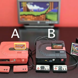 May include: Two miniature retro game consoles, one coral and one black, with matching controllers and game cartridges. A television displays a pixelated video game scene in the background. The consoles are labeled A and B.