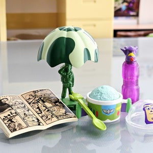 Puede incluir: Un juguete de paraca&iacute;das de camuflaje verde con una figura de soldado, un contenedor de helado verde y blanco con una cuchara, una botella de agua morada con un personaje morado en la parte superior, un c&oacute;mic y una c&uacute;pula de pl&aacute;stico transparente con un personaje morado en la parte superior.