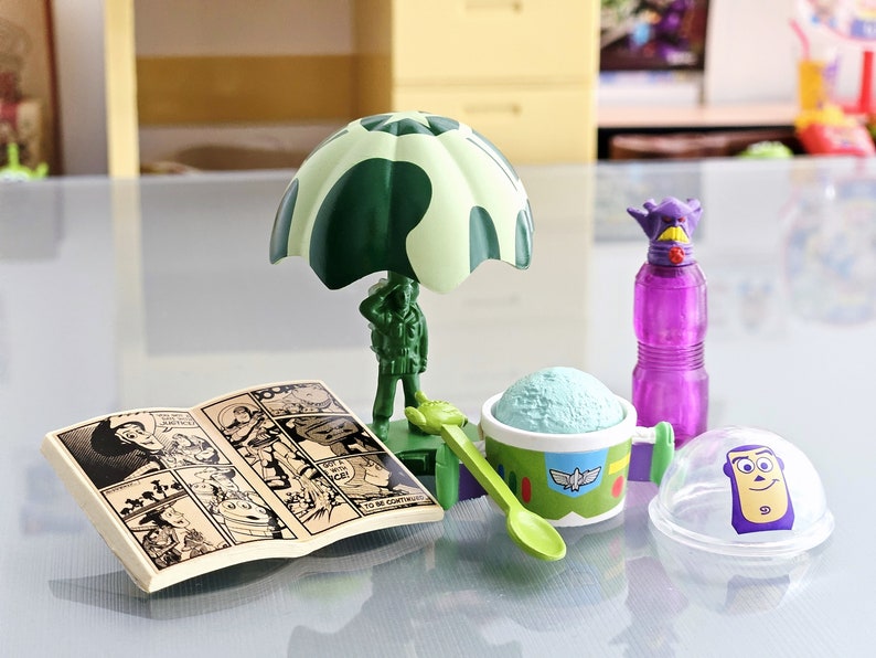 Puede incluir: Un juguete de paraca&iacute;das de camuflaje verde con una figura de soldado, un contenedor de helado verde y blanco con una cuchara, una botella de agua morada con un personaje morado en la parte superior, un c&oacute;mic y una c&uacute;pula de pl&aacute;stico transparente con un personaje morado en la parte superior.