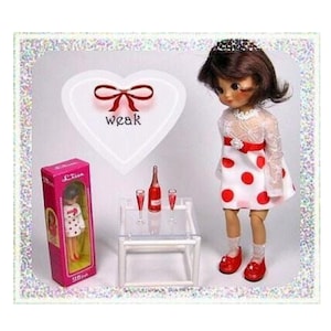 May include: A doll wearing a white dress with red polka dots, red shoes, and a tiara. A pink box with a doll inside is on the left. A bottle of red liquid and two glasses are on a small table. The word "weak" is on a heart-shaped background.