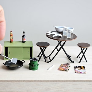 May include: A miniature dining set with a brown folding table, two black folding chairs, two white bowls, and two clear glasses. There are two bottles of sauce, a green metal container, a black wok, and a silver spatula on a green cabinet. The set is on a light wood surface.
