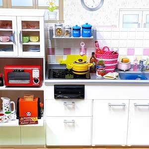 Puede incluir: Un juego de cocina de casa de muñecas blanca con un horno rojo, una tostadora, un armario blanco con puertas de vidrio y un fregadero blanco con un grifo plateado. La cocina también incluye una variedad de accesorios, como una tabla de cortar, una olla, una lata de salsa de tomate y una cesta de fruta.