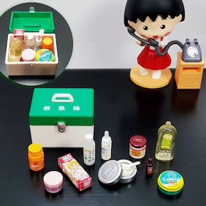 Pode incluir: Um kit de primeiros socorros verde e branco em miniatura com caracteres japoneses. Vários frascos e recipientes pequenos estão dispostos ao redor, incluindo uma lata amarela e azul com a etiqueta "Catmix". Uma figura de uma menina segurando um telefone está no fundo.