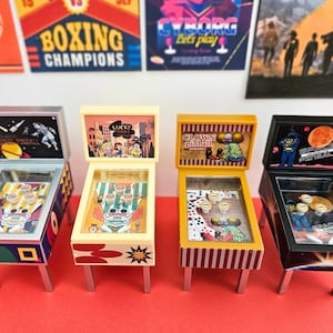 Könnte beinhalten: Vier Miniatur-Flipperautomaten mit unterschiedlichen Themen. Die Automaten sind in verschiedenen Farben gehalten, darunter Schwarz, Gelb und Creme. Jede Maschine hat ein einzigartiges Backboard-Design und ein glasbedecktes Spielfeld. Die Automaten stehen auf silbernen Beinen, mit Retro-Postern im Hintergrund.