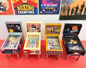May include: Four miniature pinball machines with different themes. The machines are in various colors, including black, yellow, and cream. Each machine has a unique backboard design and a glass-covered playfield. The machines are on silver legs, with retro posters in the background.