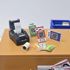 May include: A miniature black receipt printer with a roll of white paper, alongside various payment cards and QR code displays. The scene is set on a brown surface, suggesting a point-of-sale theme.