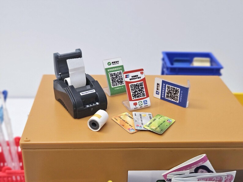 May include: A miniature black receipt printer with a roll of white paper, alongside various payment cards and QR code displays. The scene is set on a brown surface, suggesting a point-of-sale theme.