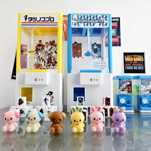Könnte beinhalten: Ein Set aus vier Miniatur-Greifautomaten mit bunten Designs und einer Auswahl an kleinen Plüschtieren. Die Greifautomaten sind mit "Crane game" und "タツノコプロ" beschriftet.