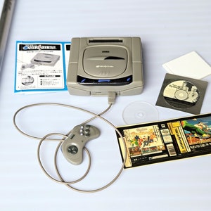 May include: A gray Sega Saturn video game console with a controller, a game cartridge, and a CD. The console has a black screen with the Sega logo. The controller is connected to the console with a gray cable. The game cartridge is inserted into the console. The CD is in a black case with a white label.
