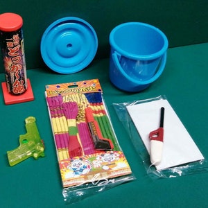 Könnte beinhalten: Ein Set Partyzubehör, bestehend aus einem blauen Eimer, einem rot-weißen Feuerwerkskörper, einer grünen Spielzeugpistole, einer Packung bunter Papier-Serpentinen, einem weißen Blatt Papier und einem rot-weißen Feuerzeug.