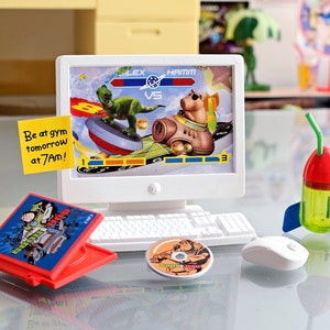 Puede incluir: Un conjunto de ordenador de juguete con un monitor que muestra un videojuego con un dinosaurio y un personaje de cerdo. El juego se titula "Lex vs Hamm". Hay una caja de CD de juguete roja con una imagen de dibujos animados de un personaje en la portada. Una nota adhesiva amarilla dice "Be at gym tomorrow at 7am!" Hay una botella de agua de juguete con forma de cohete con una pajita verde y un rat&oacute;n de juguete blanco.