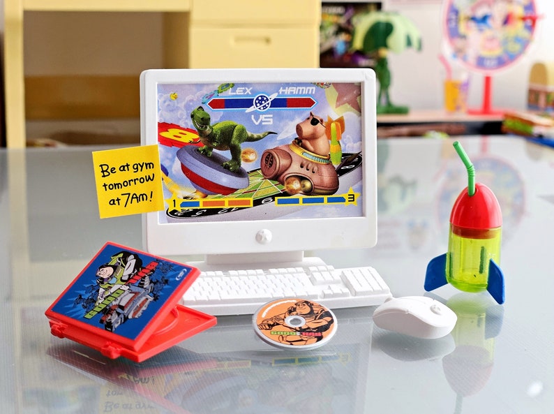 Puede incluir: Un conjunto de ordenador de juguete con un monitor que muestra un videojuego con un dinosaurio y un personaje de cerdo. El juego se titula "Lex vs Hamm". Hay una caja de CD de juguete roja con una imagen de dibujos animados de un personaje en la portada. Una nota adhesiva amarilla dice "Be at gym tomorrow at 7am!" Hay una botella de agua de juguete con forma de cohete con una pajita verde y un rat&oacute;n de juguete blanco.