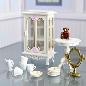 Op de afbeelding: Een witte poppenhuis-meubelset met een vitrinekast, een kleine tafel met een teddybeer erop, een spiegel en een theeservies met een theepot, kopjes en schoteltjes.