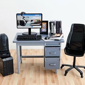 Puede incluir: Un escritorio gris en miniatura con dos cajones, un monitor de computadora negro que muestra un navegador web, un teclado negro, un ratón, una silla de oficina negra, una torre de computadora negra, un pequeño cuadro de un perro y libros.