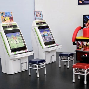 May include: Two white arcade cabinets with green screens, one displaying the game "Toys Harrier" and the other displaying the game "Ready", with three silver stools in front of them.