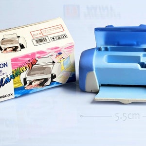 Op de afbeelding: Een blauwe en witte speelgoedprinter met een doorzichtig open deksel. De printer is 5,5 cm breed. De doos voor de printer is wit met roze en blauwe accenten en zegt "Puchison Calardaiya" in het Japans.