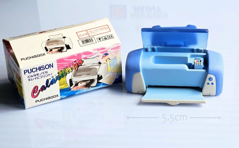 Op de afbeelding: Een blauwe en witte speelgoedprinter met een doorzichtig open deksel. De printer is 5,5 cm breed. De doos voor de printer is wit met roze en blauwe accenten en zegt "Puchison Calardaiya" in het Japans.
