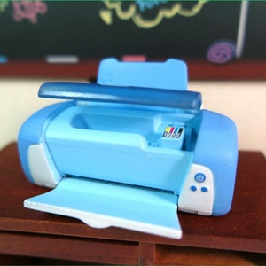 Op de afbeelding: Een lichtblauwe speelgoedprinter met een witte voorkant en een doorzichtig deksel. De printer heeft een papierlade en een knop aan de voorkant. De printer staat op een houten tafel.