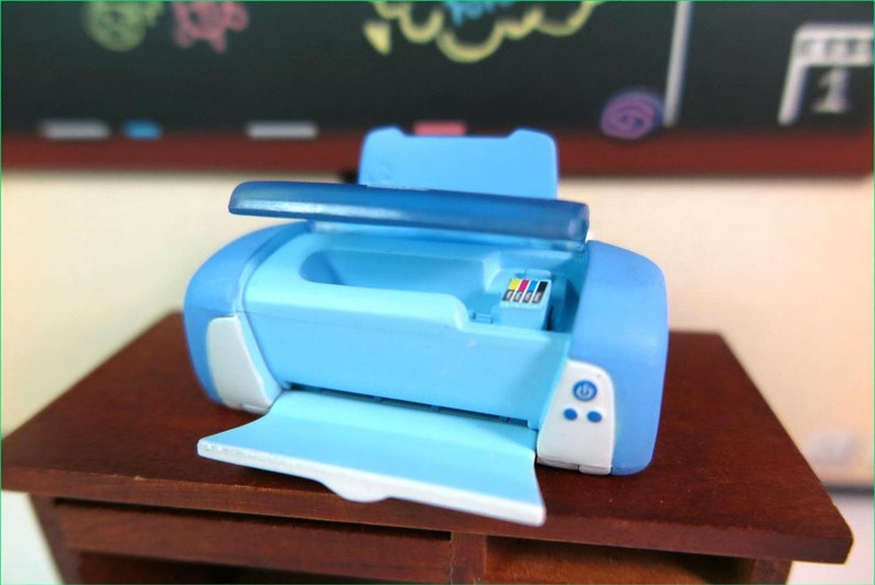 Op de afbeelding: Een lichtblauwe speelgoedprinter met een witte voorkant en een doorzichtig deksel. De printer heeft een papierlade en een knop aan de voorkant. De printer staat op een houten tafel.