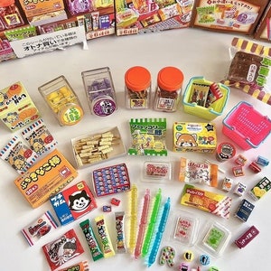 Peut inclure: Une collection d'aliments miniatures, comprenant des bonbons, des en-cas et des produits de boulangerie, disposés sur une surface blanche. Les articles sont emballés dans des emballages et des contenants colorés, et certains sont présentés dans de petits paniers de magasin. Le texte "おっきなこ棒" est visible sur l'un des emballages.