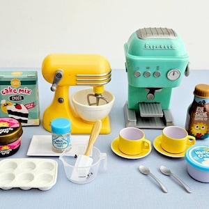 Puede incluir: Un juego de cocina de juguete que incluye una batidora de pie amarilla, una máquina de café espresso color verde azulado, una caja de mezcla para pastel, jarabe de chocolate, chispas de chocolate blanco, un molde para muffins, una taza medidora con espátula, dos tazas amarillas sobre platillos, dos cucharas y crema batida.