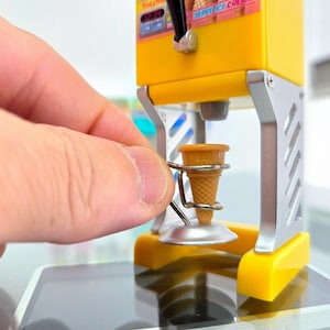 Può includere: Una macchina per gelato in miniatura di colore giallo e argento con un portacono. La macchina per gelato ha una piccola finestra con il testo "Vanilla Ice Cream" visibile. Una mano tiene il portacono.