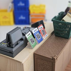 May include: Miniature cash register with a display screen, alongside three QR code payment stands. A green shopping basket holds small bottles and a block. The scene is set on a wooden counter, with blurred crates in the background.