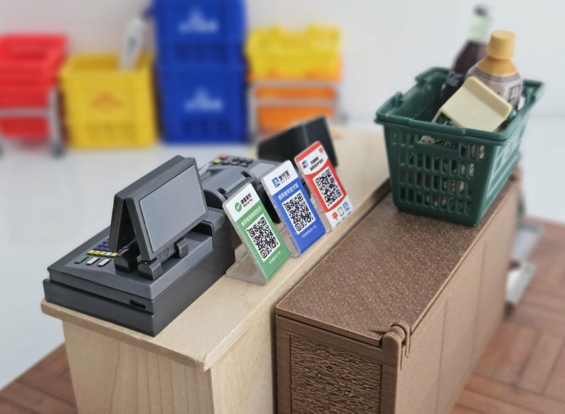 May include: Miniature cash register with a display screen, alongside three QR code payment stands. A green shopping basket holds small bottles and a block. The scene is set on a wooden counter, with blurred crates in the background.