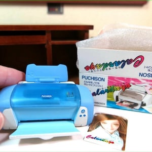 Op de afbeelding: Een blauwe en witte speelgoedprinter met een transparant plastic deksel. De printer is gelabeld met "Puchison" en heeft een open papierlade. De printer staat op een houten oppervlak. De doos voor de printer staat op de achtergrond en is gelabeld met "Calarday" en "Puchison".