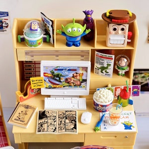 Puede incluir: Un escritorio amarillo de juguete con una computadora, una consola de juegos de juguete, un libro de juguete, un c&oacute;mic de juguete, un cubo de palomitas de ma&iacute;z de juguete, una bebida de juguete y un personaje de juguete de la pel&iacute;cula Toy Story. El escritorio est&aacute; decorado con personajes de juguete de la pel&iacute;cula Toy Story.
