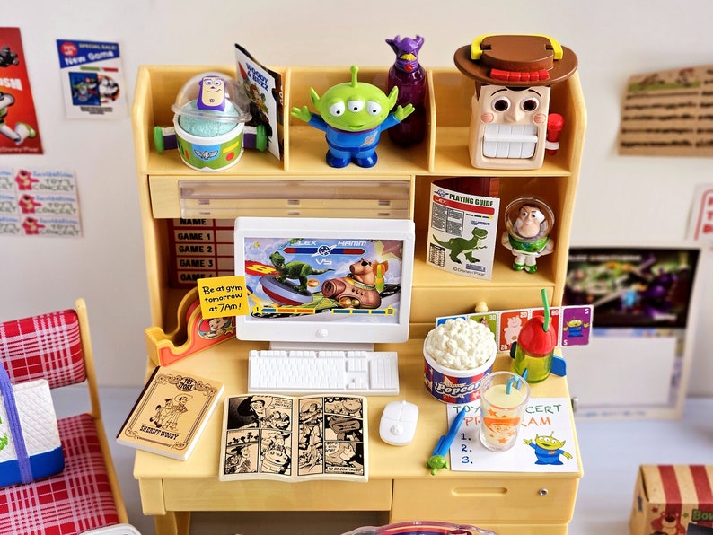 Puede incluir: Un escritorio amarillo de juguete con una computadora, una consola de juegos de juguete, un libro de juguete, un c&oacute;mic de juguete, un cubo de palomitas de ma&iacute;z de juguete, una bebida de juguete y un personaje de juguete de la pel&iacute;cula Toy Story. El escritorio est&aacute; decorado con personajes de juguete de la pel&iacute;cula Toy Story.