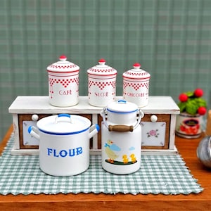 May include: A miniature kitchen scene featuring white ceramic canisters with red checkered patterns and labels for "Cafe", "Sucre", and "Chicoree". A white pot with blue trim and "Flour" text, and a milk jug with a duck and ducklings are also present.