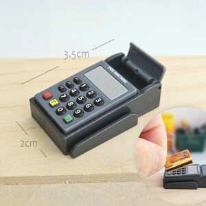 May include: A miniature gray POS machine with a keypad, a screen, and a card slot. The machine is 3.5 cm long and 2 cm wide. The keypad has black, red, yellow, and green buttons. The text "POS MACHINE" is visible on the device.