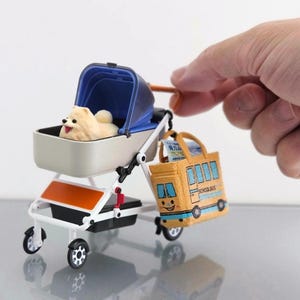 May include: A miniature toy stroller with a blue canopy and a beige dog inside. Attached is a yellow school bus-shaped bag. The stroller has a white frame with black wheels and an orange accent.