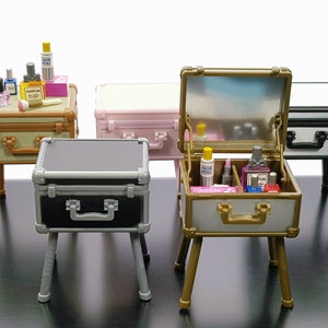 Puede incluir: Cinco maletas en miniatura con patas, cada una con un color y diseño diferente. Algunas maletas están abiertas y contienen botellas en miniatura de perfume y otros productos de belleza. Las maletas están sobre una superficie negra.
