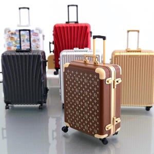 Può includere: Una collezione di cinque valigie con ruote in miniatura di vari colori, tra cui marrone con un motivo a monogramma ripetuto, oro metallizzato, argento, rosso e nero, ciascuna dotata di manico retrattile, quattro ruote e guscio rigido.